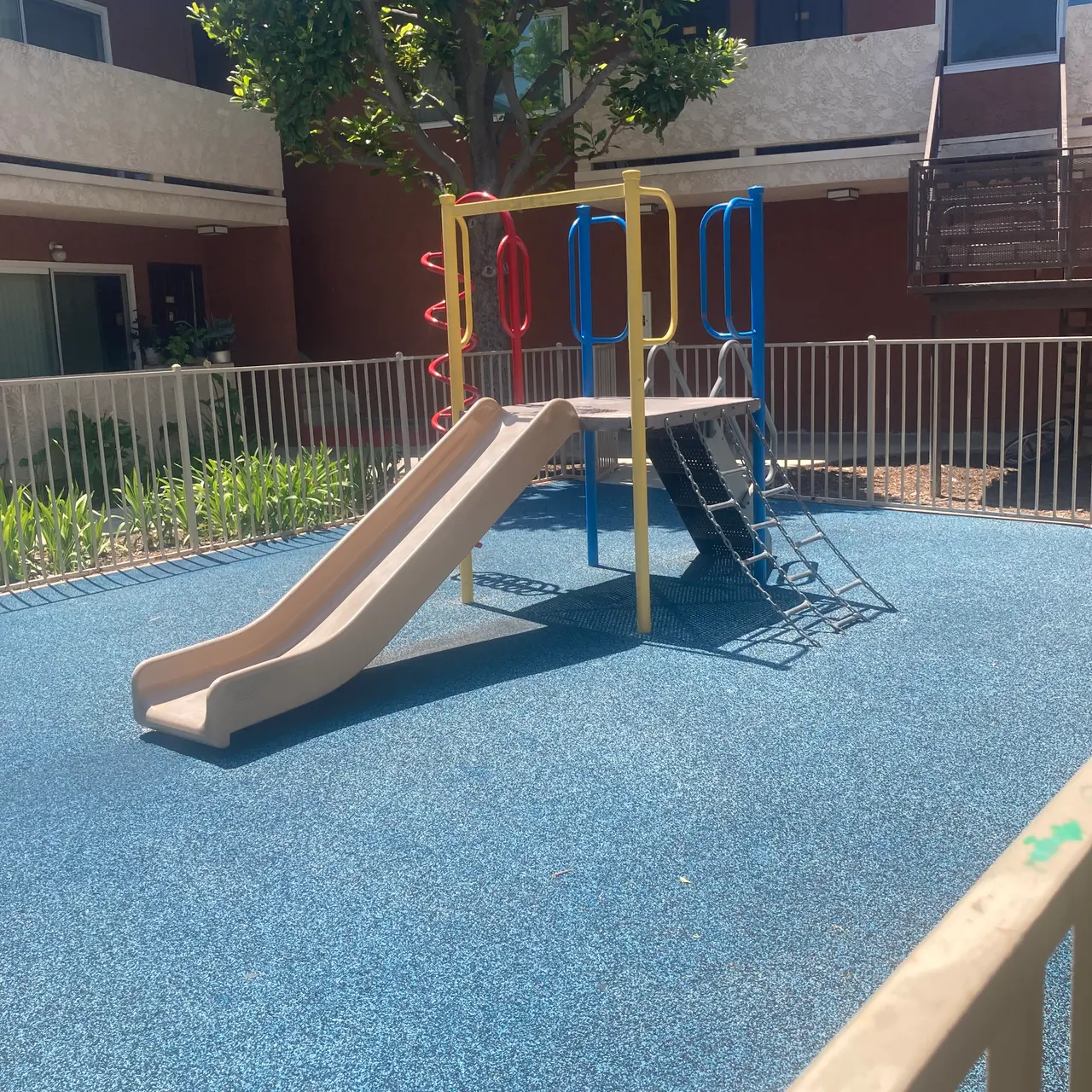A playground with a slide and climbing structure surrounded by a fence and greenery, set in a courtyard of an apartment complex.
