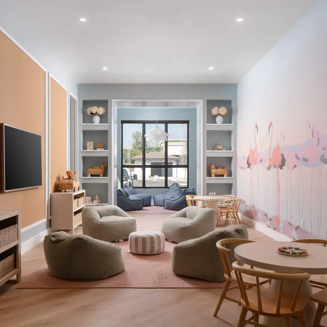 A well-designed playroom featuring soft seating, a small table with chairs, and shelves with toys. The walls are light blue with a splash of colorful mural and there is a large window letting in natural light.