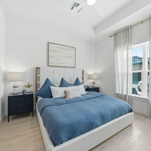 Modern bedroom featuring a white bed with blue bedding, nightstands with lamps, and large windows with sheer curtains.