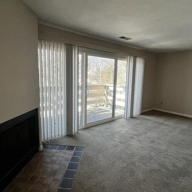 An empty living room with beige walls and carpet, featuring a fireplace on the left and large windows with sheer curtains leading to a balcony.