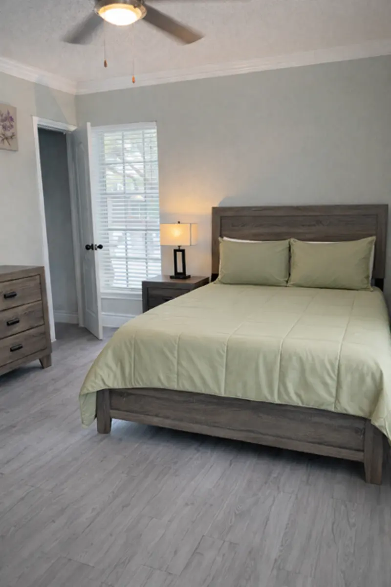 A serene bedroom featuring a wooden bed with a light green bedspread, two matching pillows, a bedside table with a lamp, and a dresser. Windows with white shutters provide natural light, and a ceiling fan is visible overhead.