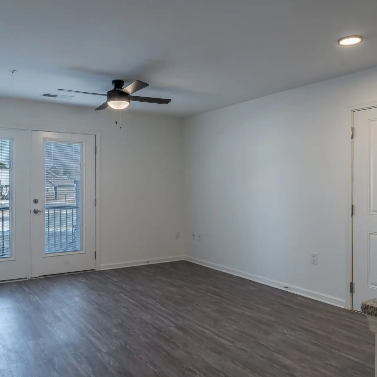 A spacious living room with light gray walls, a ceiling fan, and wood-like flooring. Two doors leading to a balcony and a door to another room are visible. The room is bright with natural light.