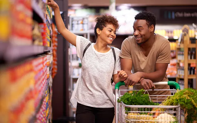 A young couple enjoying their time while grocery shopping in a colorful supermarket aisle. The woman, with curly hair and a striped shirt, is smiling and looking at the man, who is wearing a tan shirt. They are next to a shopping cart filled with fresh produce.