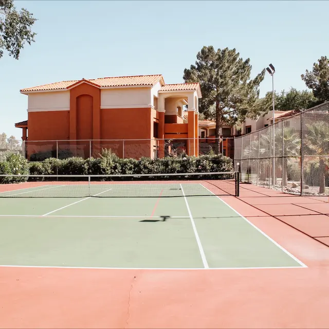 View of a green tennis court with a fence, surrounded by residential buildings and palm trees in the background.
