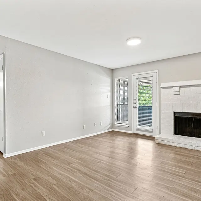 A spacious and modern living room with wood flooring. There are white walls and a white brick fireplace on the right. On the left side, there are two closed doors leading to other rooms. Large sliding glass doors open to a small balcony or patio area.