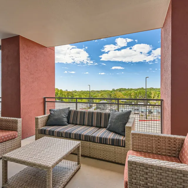 An outdoor seating area with two armchairs and a couch on a balcony. The furniture has beige wicker design, with cushions in shades of orange and gray. A coffee table is in the center. In the background, there is a view of trees and a blue sky with fluffy white clouds.