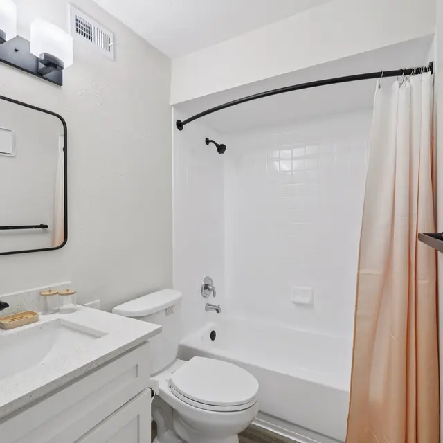 A modern bathroom featuring a white bathtub with a shower curtain, a rectangular mirror above a sleek vanity with a white countertop, black fixtures, and a light fixture above the mirror.