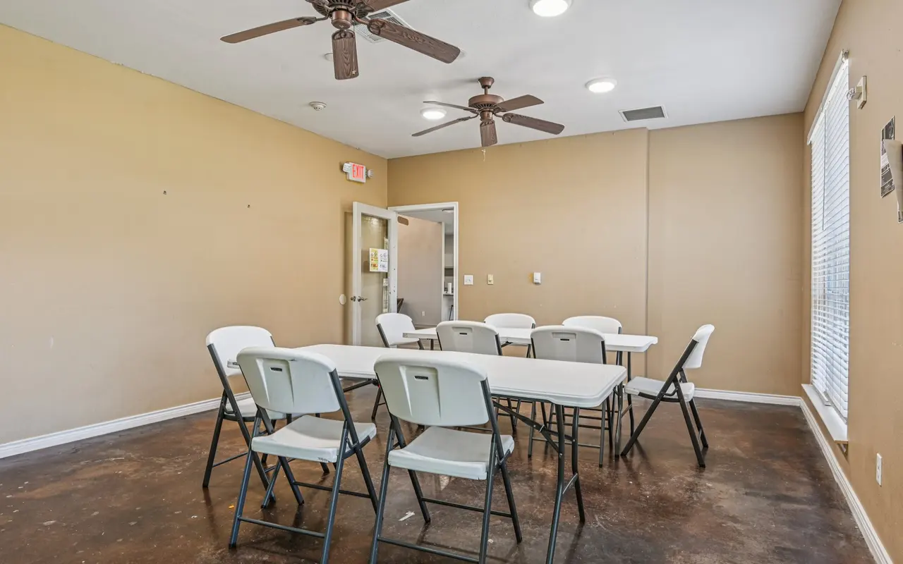 Interior of a meeting room with folding tables and chairs, two ceiling fans, beige walls, and large windows