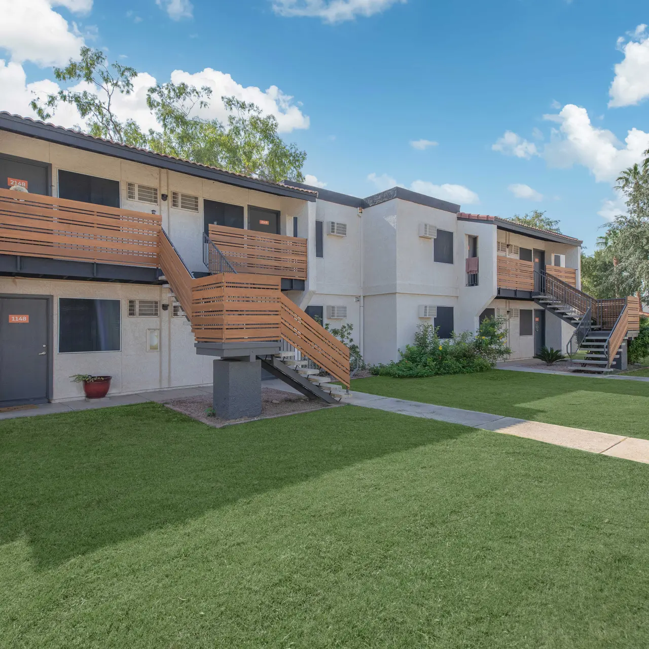 Exterior view of a two-story apartment complex with wooden staircases and green grass in the foreground.
