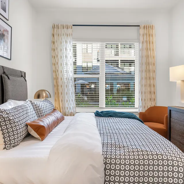 A modern bedroom featuring a comfortable bed with patterned bedding, decorative pillows, a stylish armchair, and a dresser. Large window allows natural light to brighten the space, complemented by curtain drapery.