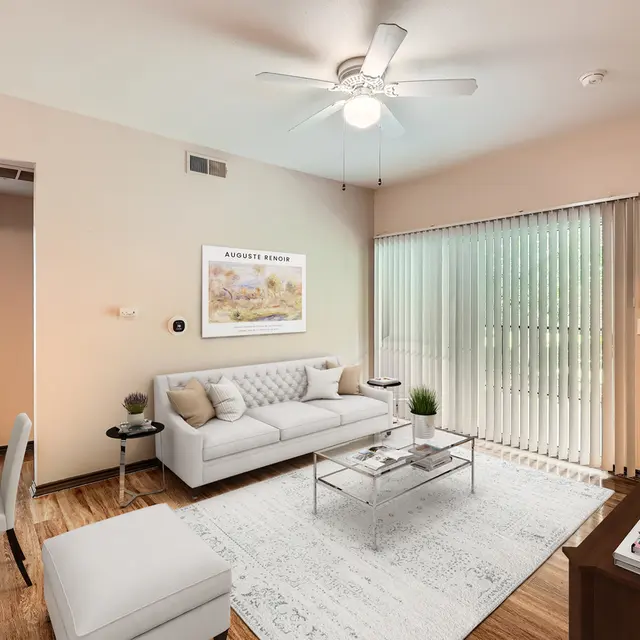 A modern living room featuring a white sofa with cushions, a glass coffee table, and a flat-screen TV. The room has wooden flooring and large sliding glass doors with vertical blinds that lead to an outdoor area. A small dining table with clear chairs is also present, and there is a wall art piece above the sofa.