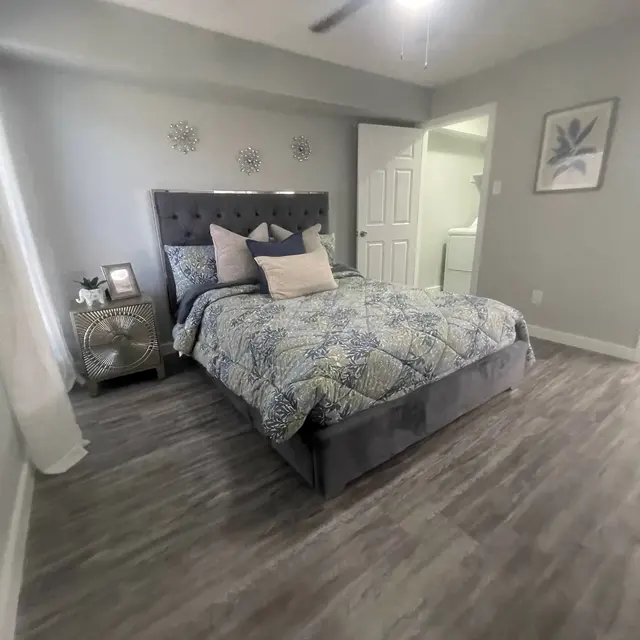 A cozy modern bedroom featuring a large bed with a tufted headboard, decorative pillows, and a colorful comforter. There is a nightstand with a lamp and decorative items. The room has light gray walls and flooring with a vent fan on the ceiling, and a door leading to another room. A framed picture is visible on the wall, suggesting a calm and stylish atmosphere.