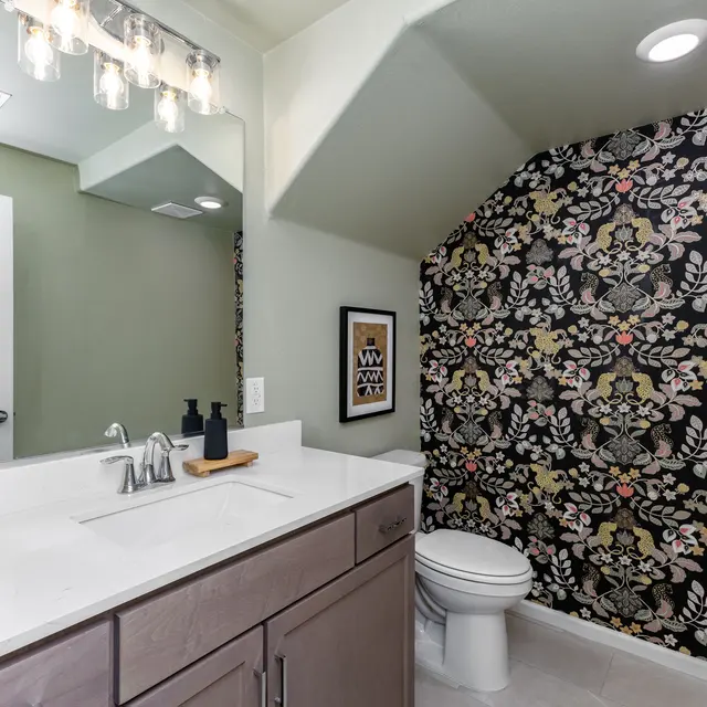 A beautifully designed bathroom featuring a modern vanity with a white countertop, a stylish black and gold patterned wallpaper on one wall, and a well-lit mirror. The space includes a white toilet and decorative items on the counter.