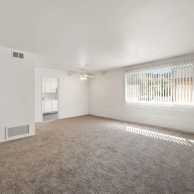 A spacious living room with a ceiling fan, a large window featuring blinds, light-colored walls, and a carpeted floor.