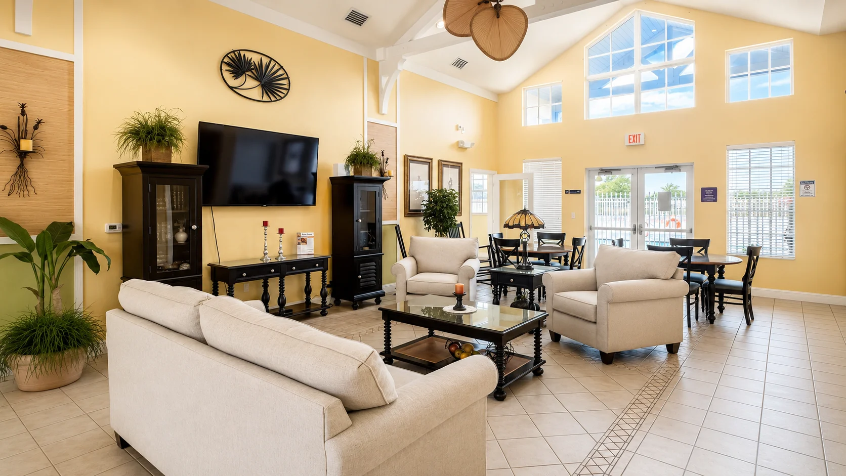 A spacious community lounge area with a large window, beige sofas, a coffee table, and a dining area.
