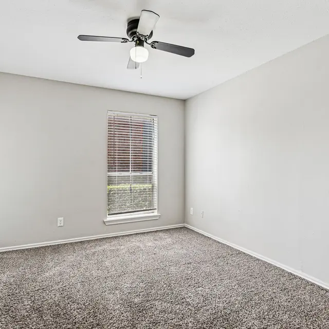 A spacious, empty bedroom with a ceiling fan and carpeted floor. There is a window with blinds on one wall, allowing natural light to enter the room.