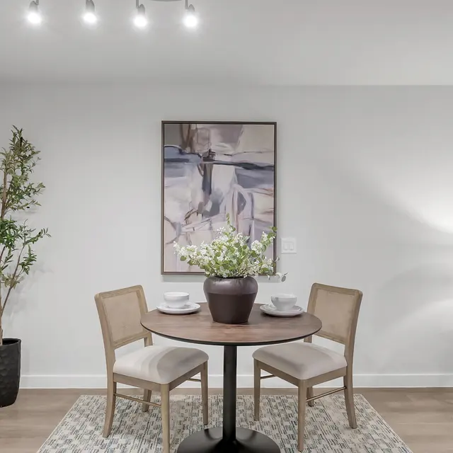 A cozy dining area with a round table and two chairs, adorned with a vase of flowers. The backdrop features a piece of abstract art and decorative circular mirrors on the wall. A potted plant stands beside the table, and a lamp casts soft light in the corner, contributing to the inviting atmosphere.