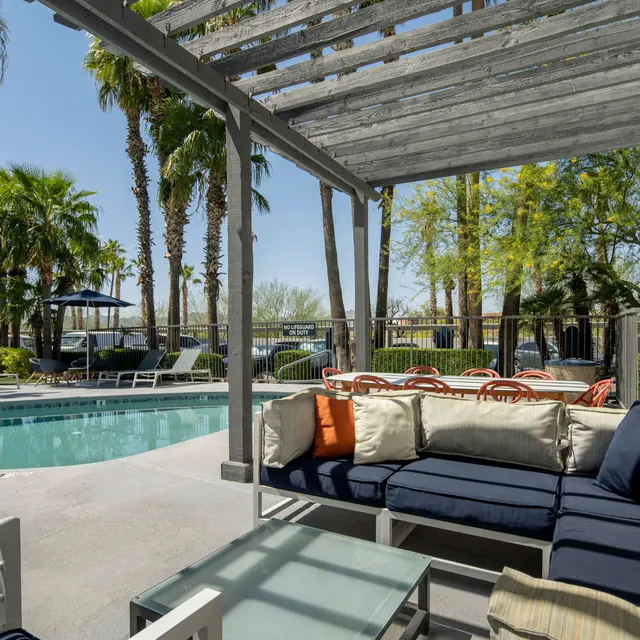 A cozy outdoor lounge area by a pool, featuring gray and navy seating with orange and cream colored pillows. Palm trees and sun loungers are visible in the background, along with a clear blue sky.