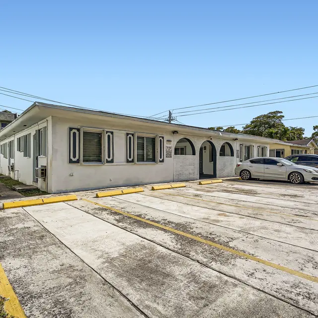 Exterior view of a single-story apartment complex with a parking lot in front and greenery nearby.