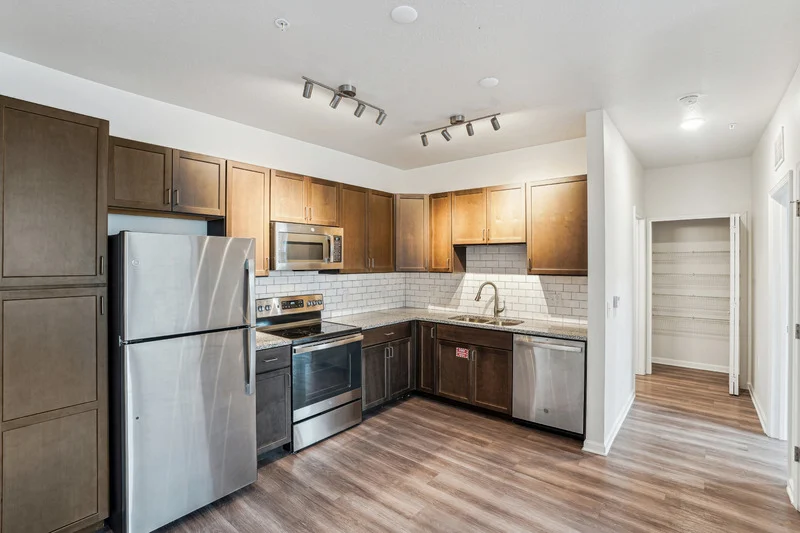 A modern kitchen featuring stainless steel appliances, including a refrigerator, oven, and dishwasher. The cabinets are dark wood with a tile backsplash. The floor has a light brown wood-like finish, and there is a doorway leading to another room.