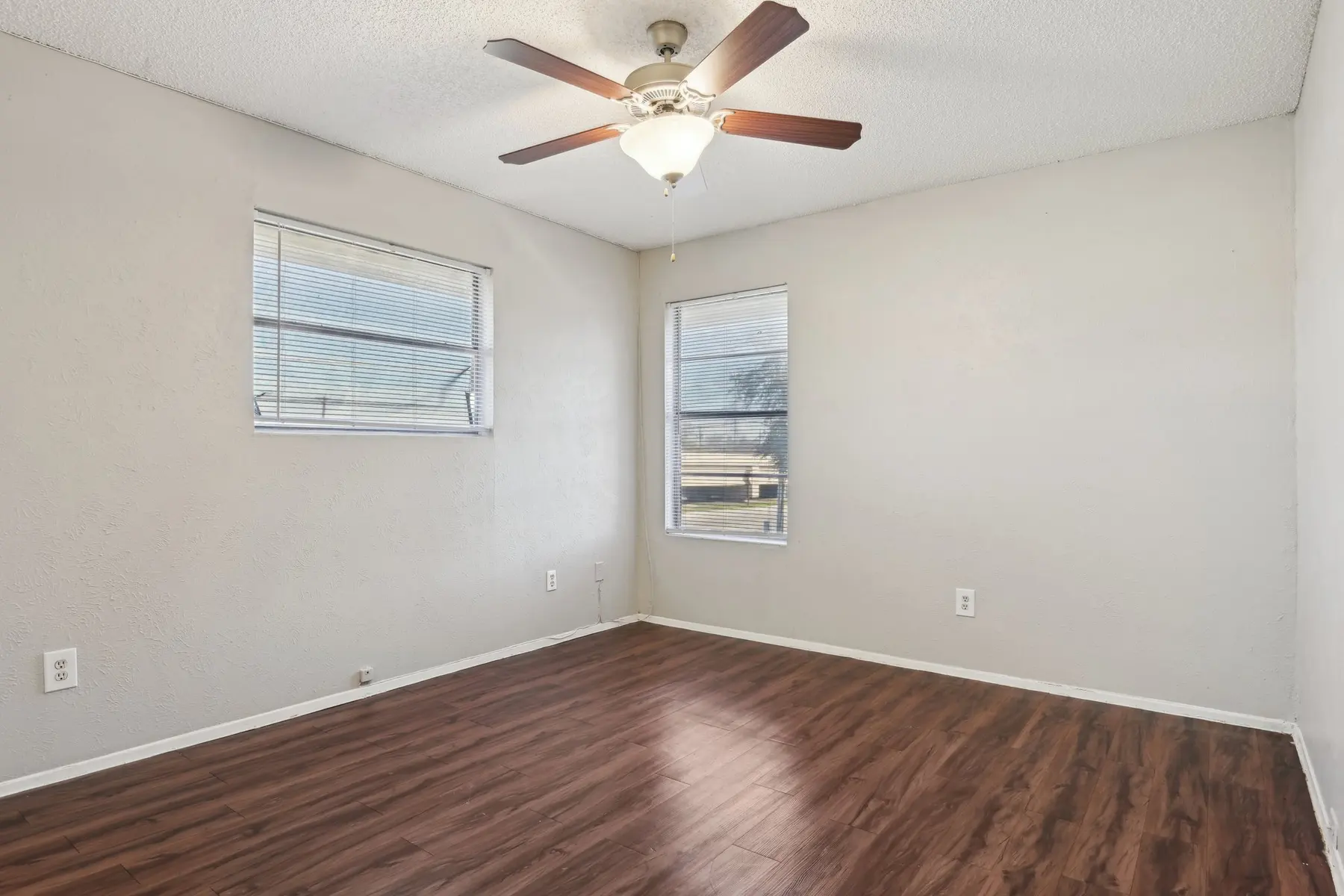 An empty bedroom with light brown wooden flooring, a ceiling fan, and two windows letting in natural light.