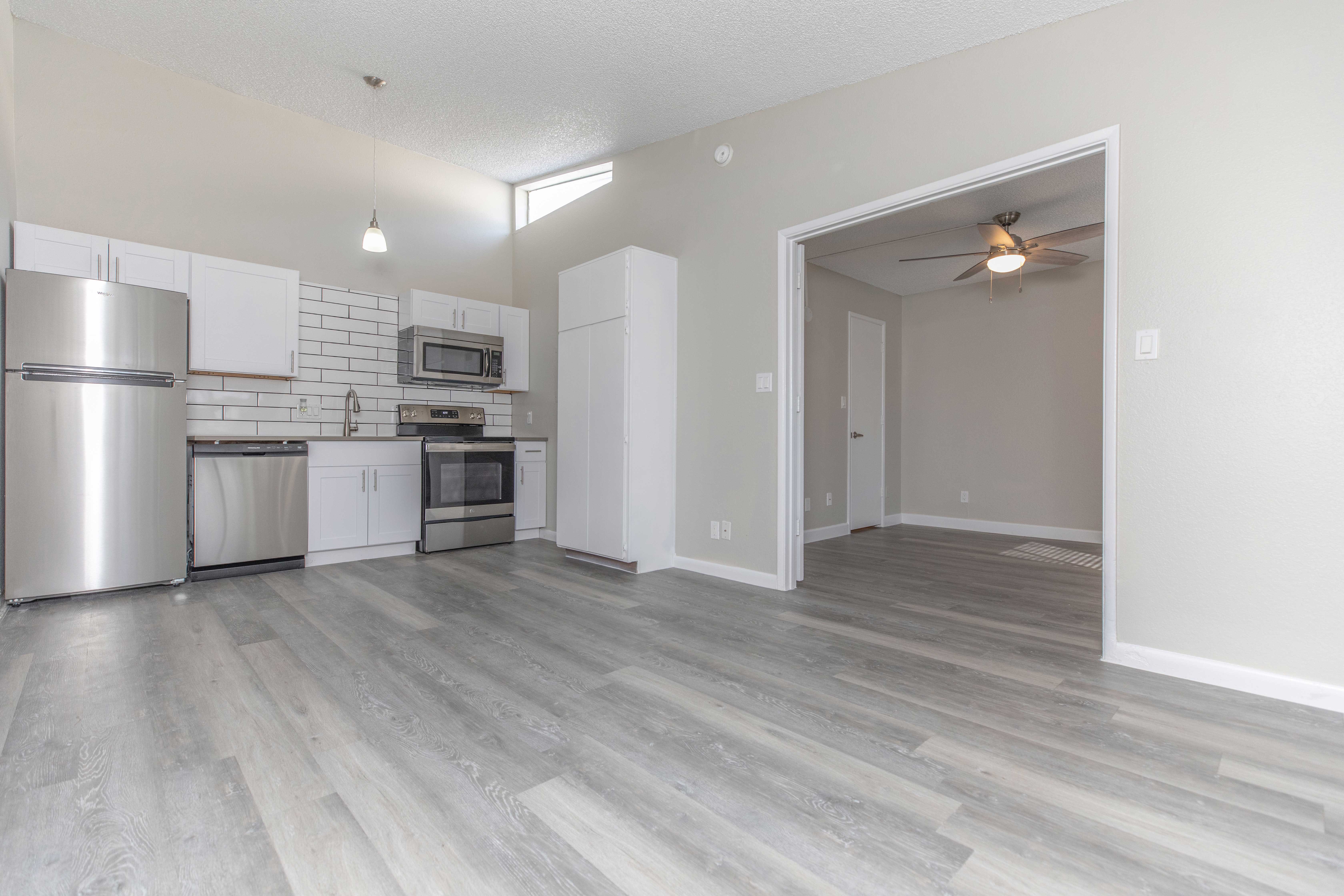 Modern Open-Concept Living Area A modern open-concept living area featuring a kitchen with stainless steel appliances, white cabinetry, and a spacious layout. Light-colored laminate flooring runs throughout the space, leading to a partially visible room with a ceiling fan.
