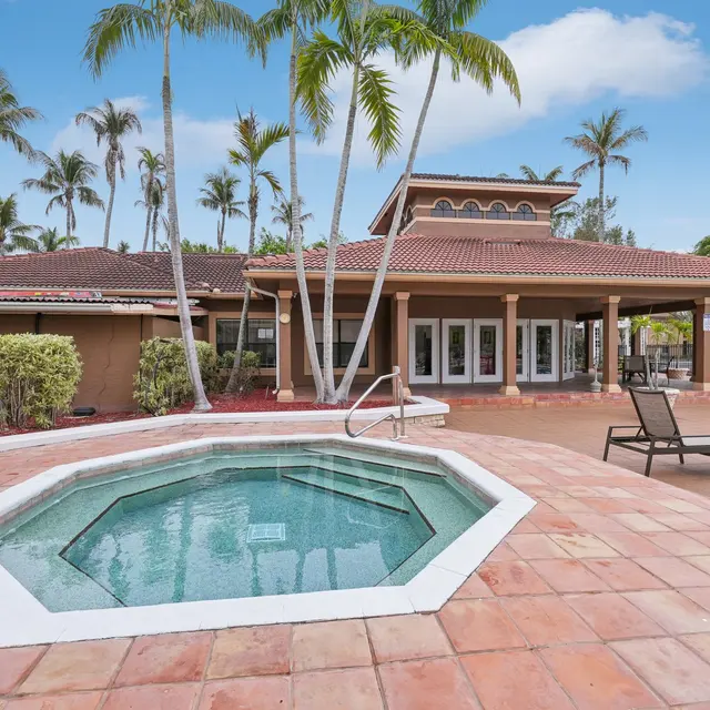A spacious backyard with a hot tub and palm trees, featuring a large patio area and a house with a tiled roof in the background.
