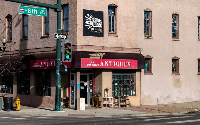 A street view of an antique store with a bright red awning labeled 'ANTIQUES'. The building has a pink exterior with large windows and a sign for the Art District. There are traffic lights and a street corner visible with a green sign indicating '8th Ave'.