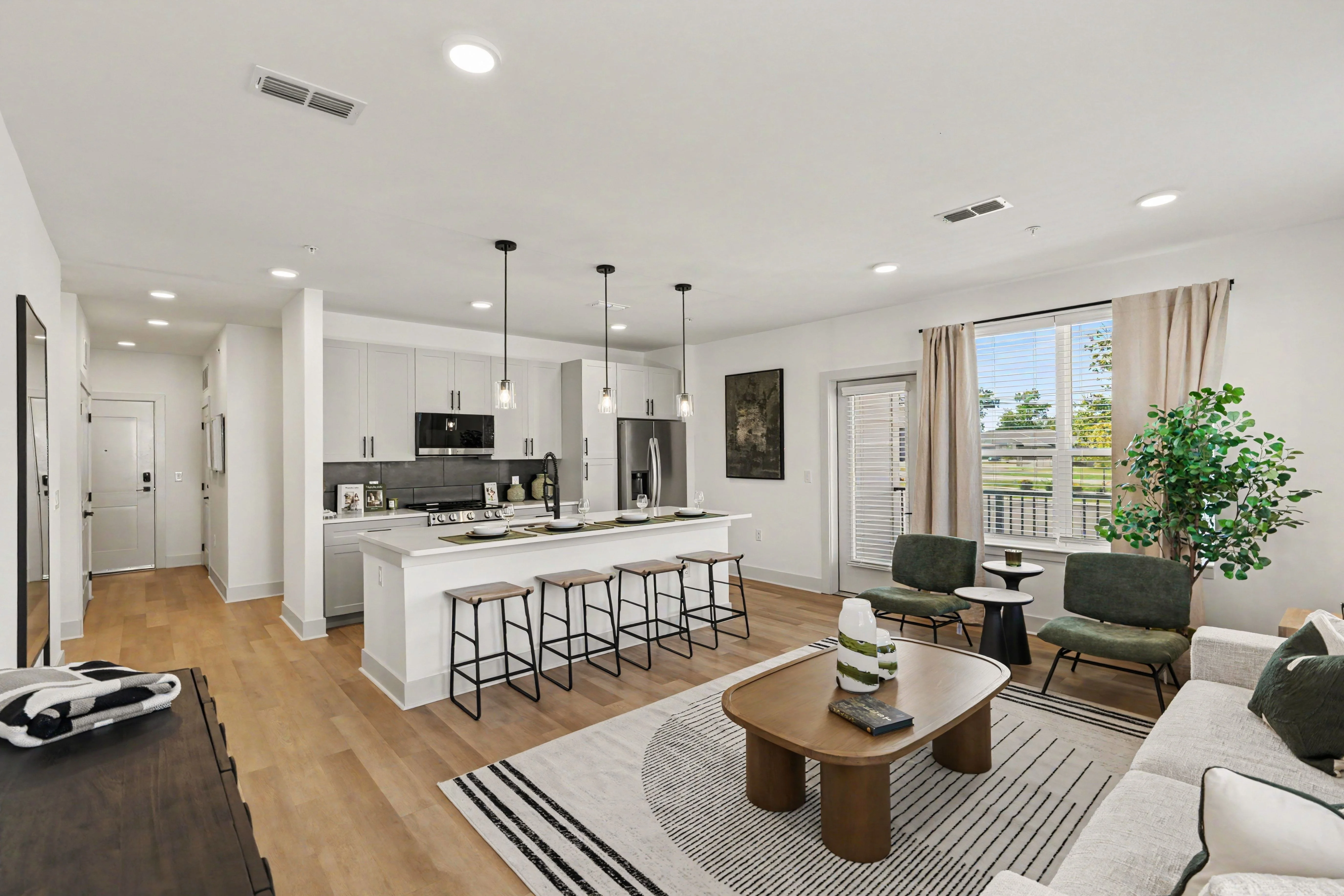 A modern open-concept living area featuring a kitchen, dining area, and living room. The kitchen has a large island with bar stools, sleek cabinetry, and stainless steel appliances. The living room includes a couch, coffee table, and green plants near a window.