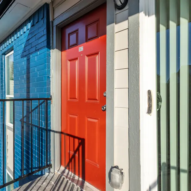 A bright red door on a multi-story building with blue painted walls. The door has the number 31 on it. A balcony railing is visible in the foreground, and there are vertical blinds on the side.