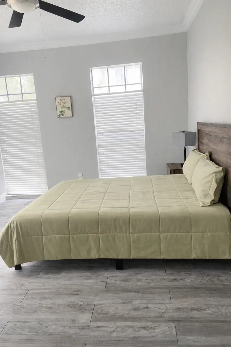 A minimalist bedroom featuring a light-colored bedspread on a bed with two pillows, placed against a wooden headboard. There are two windows with blinds and a small nightstand beside the bed.