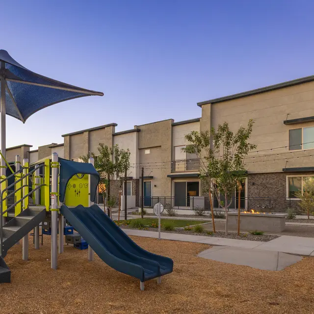 A playground featuring slides and climbing equipment, surrounded by landscaped shrubs and trees, with modern apartment buildings in the background under a clear sky.