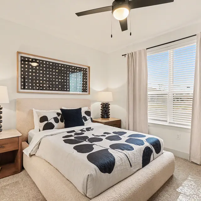 A modern bedroom featuring a bed with a patterned duvet, two bedside tables with unique lamps, a window with light curtains, and a decorative wall mirror.