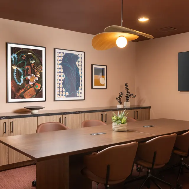 A modern meeting room featuring a long wooden table, brown chairs, and a large television mounted on the wall. Three framed artworks in varying styles are displayed on the left wall, with a light fixture above the table. A small plant is centered on the table.