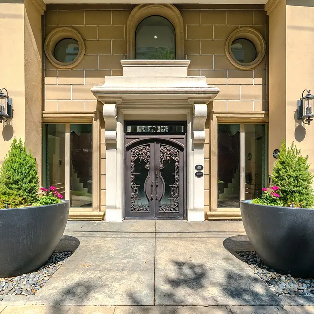 Sophisticated entrance of a luxury apartment building featuring an ornate brown door, decorative lights, and modern planters with greenery and flowers.