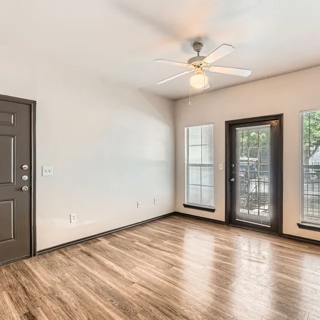 A spacious living room with a ceiling fan, wooden flooring, and large windows providing natural light.