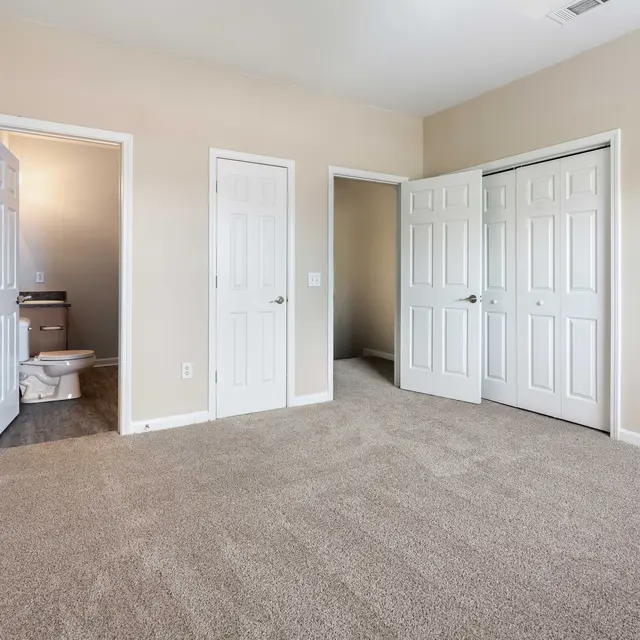 A spacious, empty room featuring beige walls and a carpeted floor. There are three doors leading to other areas. One door is ajar, revealing a glimpse of another room, while another is fully closed. The lighting is bright and inviting.