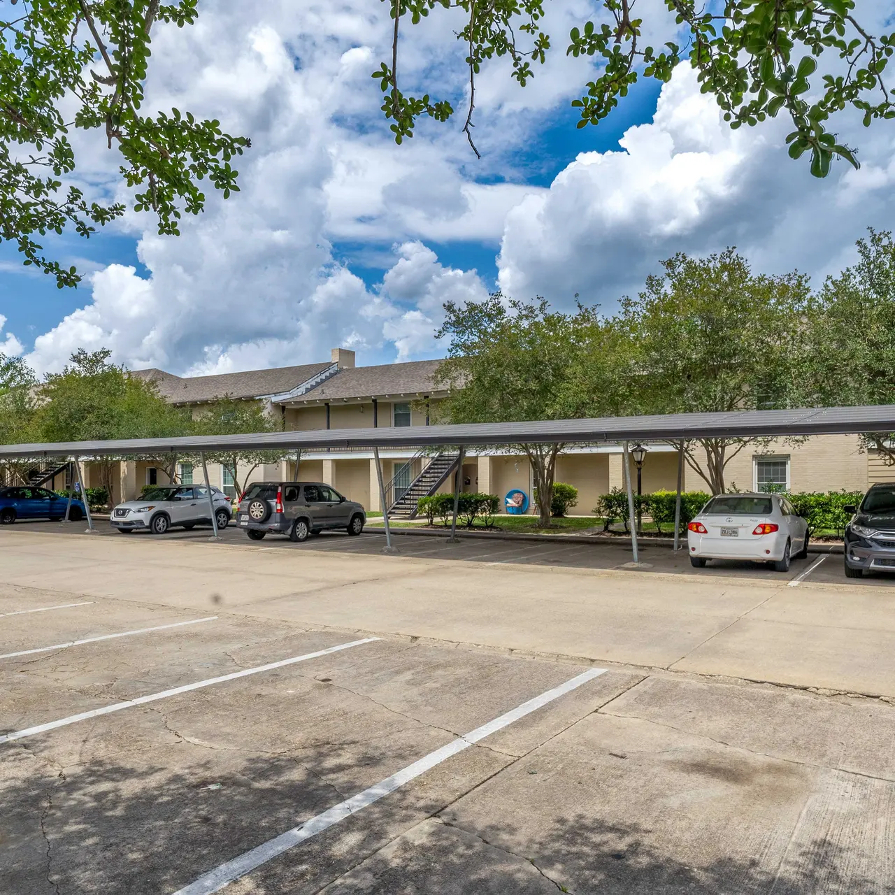 A parking lot of an apartment complex with covered spaces and several vehicles parked underneath. Cloudy blue sky with scattered clouds can be seen above.