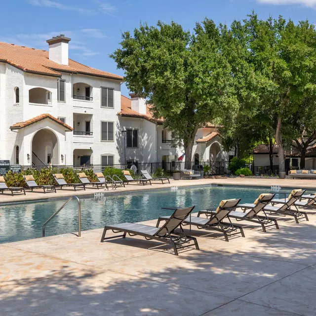 A clear swimming pool surrounded by lounge chairs and trees, with an apartment building in the background.