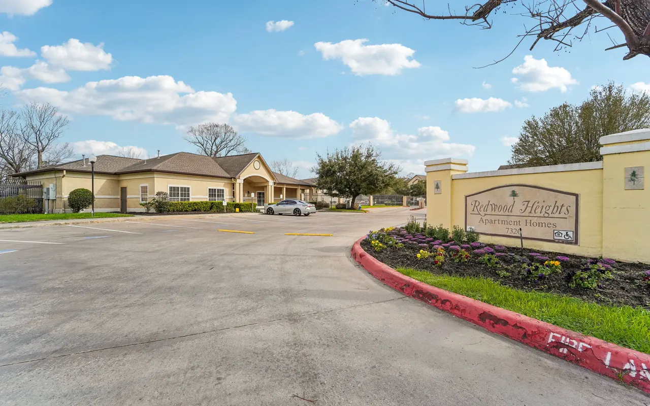 Entrance to Raymond Heights apartment complex, with a yellow sign and well-maintained landscaping under a blue sky with clouds.