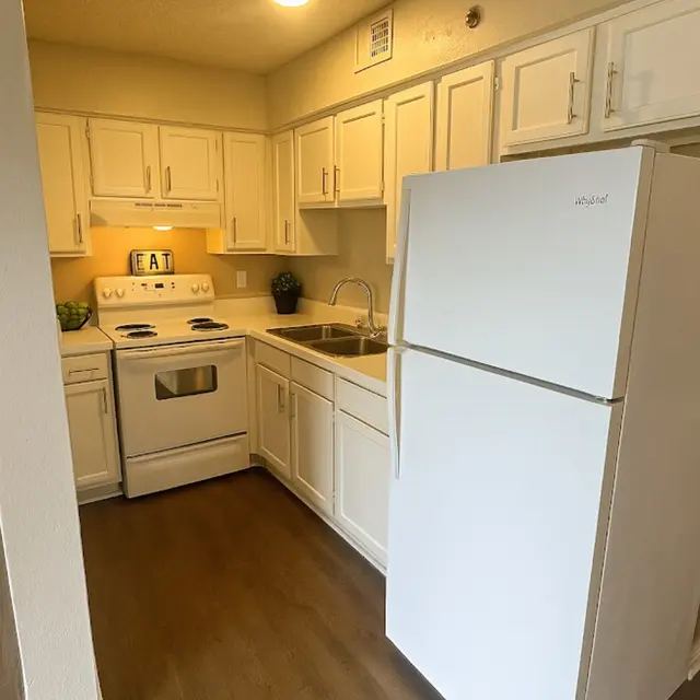 A modern kitchen featuring white cabinetry, a white refrigerator, and an oven with a stove. The kitchen has a wooden floor and is well-lit with overhead lighting.