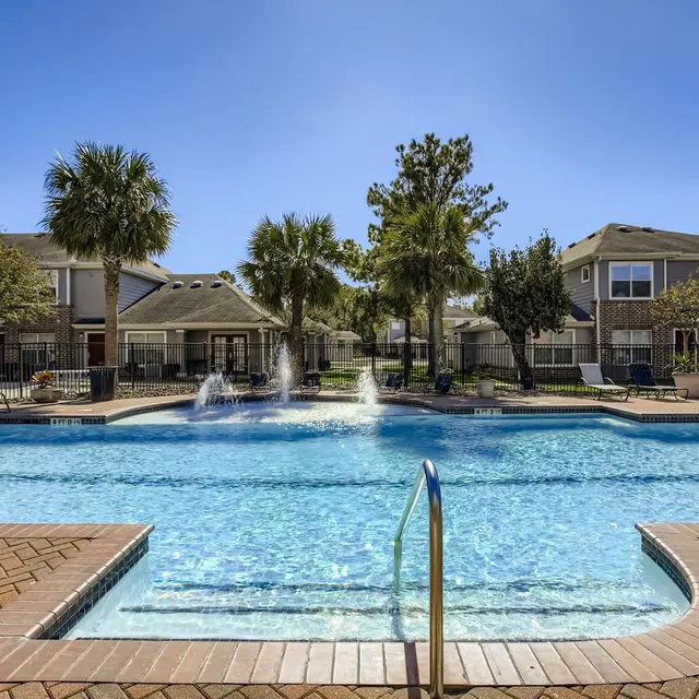 A residential pool area featuring a large swimming pool with fountains, surrounded by palm trees and well-maintained landscaping. There are several lounge chairs and a covered seating area in the background.