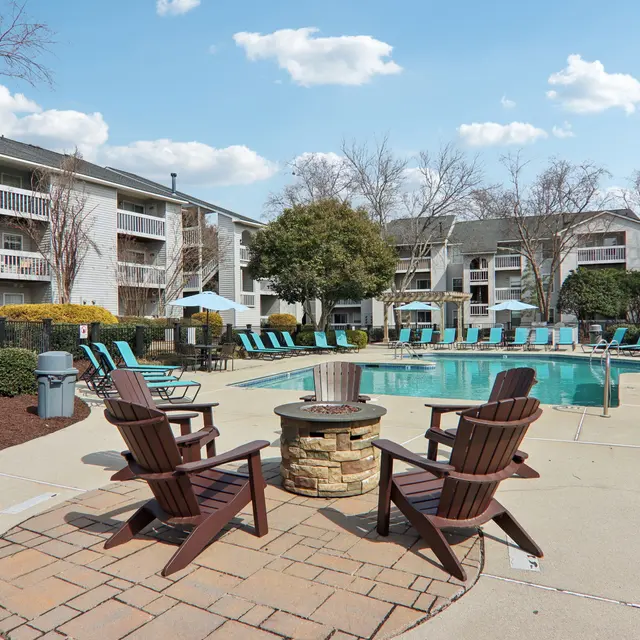A pool area in an apartment complex featuring a swimming pool surrounded by lounge chairs, a fire pit with Adirondack chairs, and nearby trees.