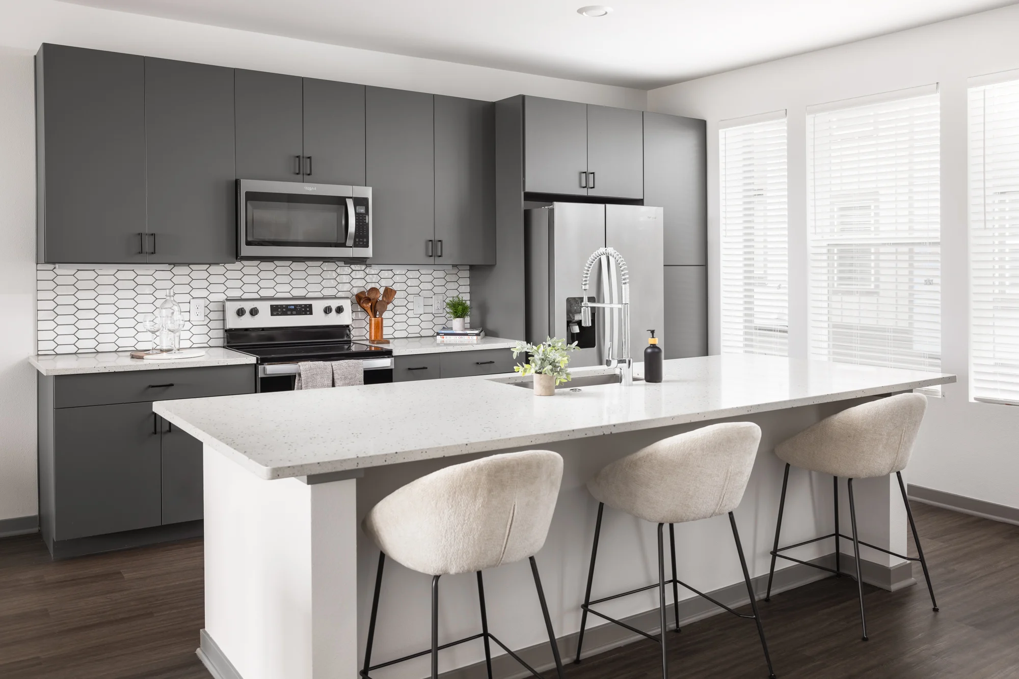 A modern kitchen with dark gray cabinets, a white countertop, stainless steel appliances, and a light backsplash. There are four beige bar stools at the counter, and large windows letting in natural light.