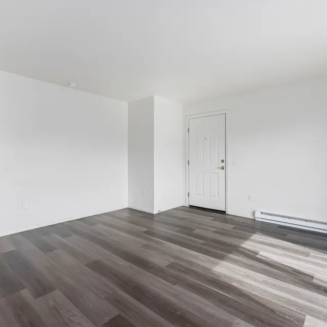 Empty Apartment Room Spacious empty room with white walls and dark wooden floors, featuring a door on one side and a window with vertical blinds on the opposite side.