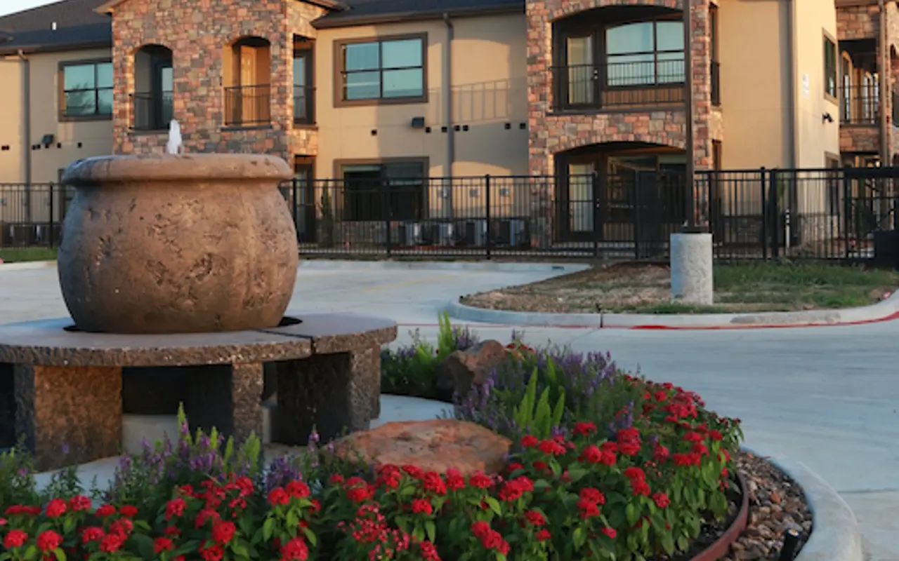 A landscaped area with a round flower bed filled with red flowers and greenery, featuring a decorative fountain, in front of a modern apartment complex.