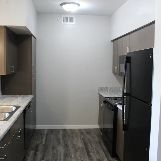 A modern kitchen featuring grey cabinets, stainless steel appliances, and a marble countertop. The kitchen has a double sink and a dark fridge, with smooth wooden flooring.