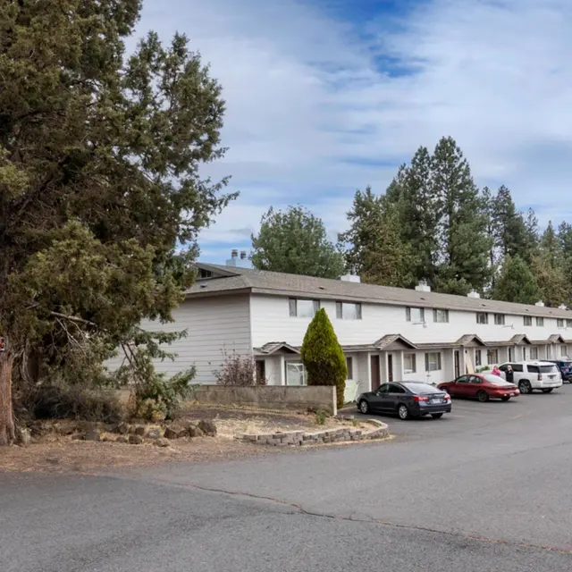 A view of a residential apartment complex on a quiet street with parked cars and trees lining the area under a partly cloudy sky.