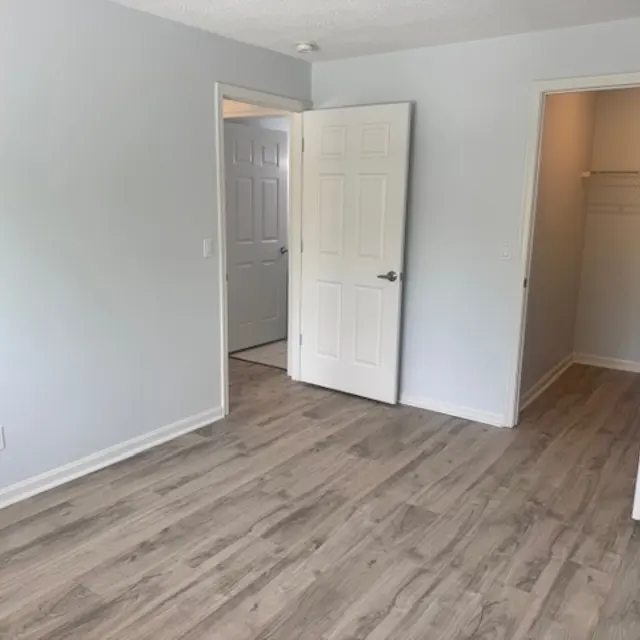 A spacious empty room with light gray walls and wooden flooring. Two doors are visible, one leading to another room and the other to a small closet.