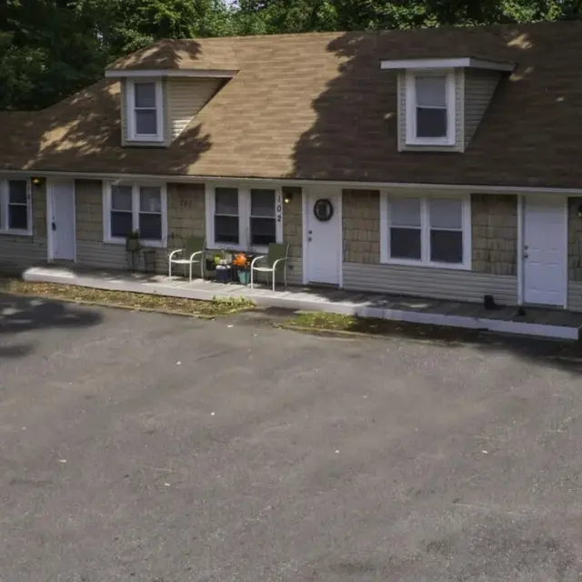 A view of a residential building with multiple units, featuring a light-colored exterior and a tan roof. The building has several windows and porch areas with seating. The surrounding area is landscaped with trees. A paved parking area is visible in front.